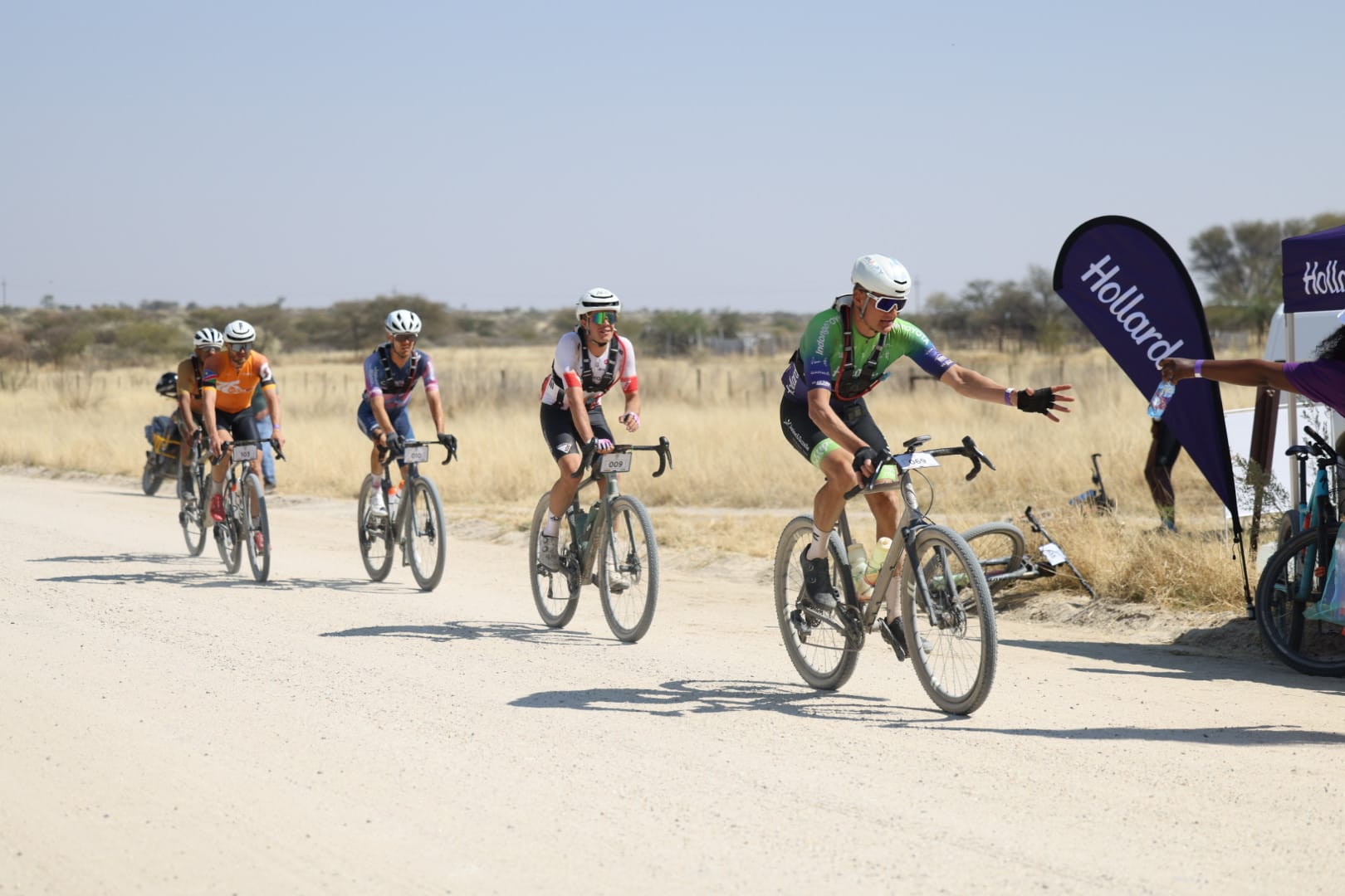 A group of cyclists competing in the Khomas100 gravel race on a dusty dirt road, with the lead rider reaching out to grab a water bottle at a Hollard-sponsored water point.